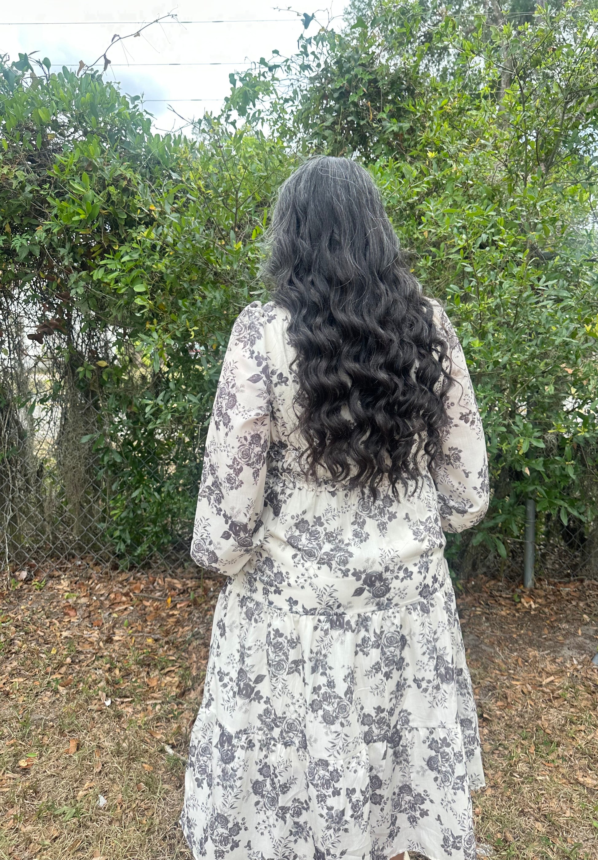 Person with long, wavy hair wearing a floral dress standing in a natural setting.