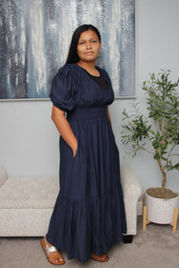 Woman wearing a navy blue dress standing in a living room with a plant and abstract painting in the background.