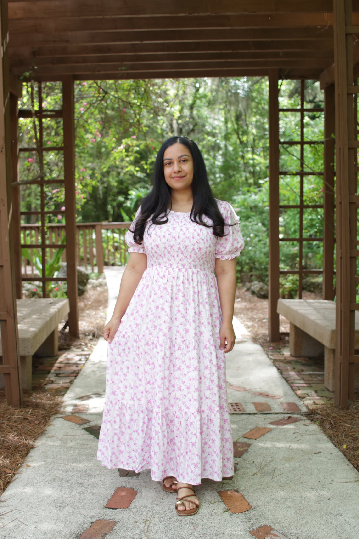 Woman in a pink dress standing under a wooden arbor with greenery in the background