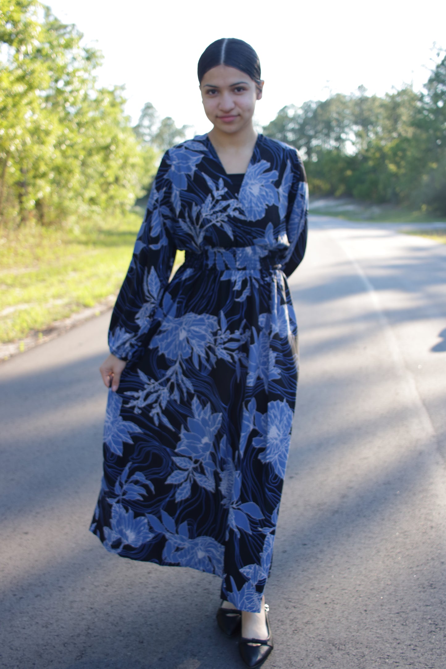 Woman wearing a blue floral dress standing on a road with trees in the background